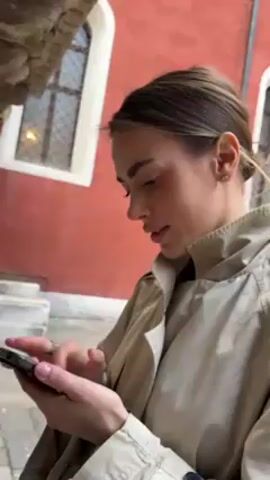 A woman is looking at her phone while standing near a red building with white-framed windows. She has brown hair pulled back, wearing a tan trench coat and small earrings. People walk behind her in the background. She appears to be focused on her phone screen.