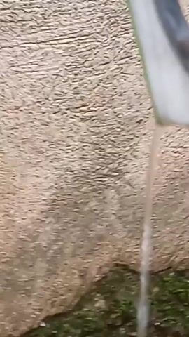 A close-up shot shows water pouring from the end of a white pipe onto a textured surface that appears to be concrete. The water stream is thin and breaks into droplets as it falls. The concrete surface is damp in places, and green moss or grass is visible at the bottom of the frame. The lighting suggests an outdoor setting.