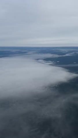 The video shows an aerial view, likely from an airplane, looking down on a layer of clouds. The sky above is overcast with gray clouds. The cloud layer below is thick and white, with areas where land and water are visible through gaps in the clouds. The overall scene is muted in color due to the cloudy conditions. There are no discernible people, objects besides clouds, or actions beyond the movement of the aircraft.