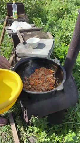 A person wearing a plaid shirt is adding water from a yellow bowl to a black pot over an outdoor stove. The pot contains cooked food with rice and carrots. The stove is made of dark metal and is surrounded by green foliage. In the background, there is a white bowl on a flat surface, a plastic bottle of oil, and a metal grill.