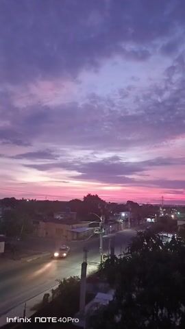 The video shows a landscape at dusk or dawn. The sky is filled with pink and purple clouds. The video is taken from a high vantage point, overlooking a neighborhood with houses and trees. A car with its headlights on is driving on a street.  The video is blurry. The camera slowly pans from left to right. The watermark "Infinix NOTE 40Pro" is visible in the lower-left corner. Someone can be heard speaking in the background.