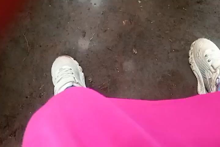 The video shows a person standing on a dirt surface. The person is wearing white sneakers and bright pink pants. The dirt surface is grey and has some debris on it. The camera is positioned looking down at the person's feet and pants.