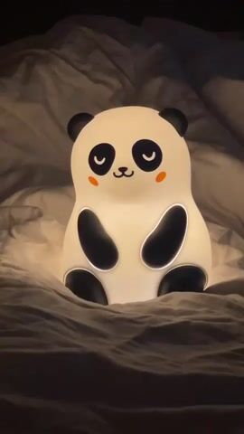 The video shows a panda-shaped night light sitting on top of a bed with white sheets. The night light is lit up, and it has black ears, black patches on its body, and a smiling face with closed eyes and rosy cheeks. The sheets are rumpled and there are some shadows on the wall behind the bed.