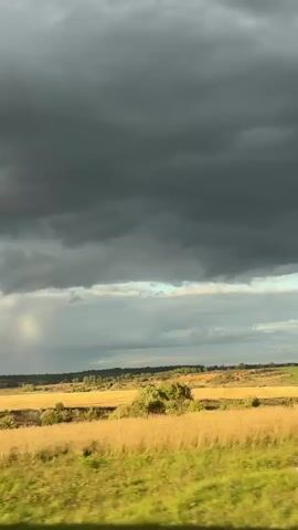 The video captures a landscape view from a moving vehicle. The sky is filled with dramatic, dark storm clouds, with lighter clouds beneath them. A sliver of sunlight breaks through on the left, creating a subtle rainbow effect. Below the sky, a vast field of dry, golden grass or wheat stretches across the horizon, dotted with small trees and bushes. The foreground shows a blurred out element of the vehicle's interior or exterior, possibly a car window or side panel. The overall scene suggests an approaching storm over a rural, agricultural area.