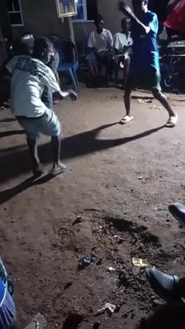 This is a low-light video of an outdoor gathering. In the foreground, two individuals are dancing. One dancer, wearing a white t-shirt with lettering and light-colored shorts, is in the center of the frame, facing left and moving their arms. Their shadow is cast prominently on the ground. To the right, another individual, wearing a blue t-shirt and dark shorts, is also dancing, facing the center. Their shadow is also visible. Behind the dancers, several other people are seated on chairs, observing the activity. The environment appears to be a dirt or gravel area, possibly a yard, with some debris on the ground. A building with a framed picture hanging on it is visible in the background. The overall atmosphere suggests a casual social event, likely a celebration or party.