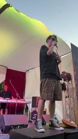 The video shows a male performer on a stage, singing into a microphone. He is wearing a black t-shirt, leopard-print shorts, and high-top sneakers. Behind him, there is a large tent-like structure with lights and sound equipment. Other people are visible in the background, some dancing. The scene appears to be an outdoor event, possibly a music festival. The performer is actively engaged with the audience, gesturing with his hands and moving his body. The lighting is dynamic, with spotlights illuminating the stage and creating a vibrant atmosphere. The overall impression is one of an energetic live performance.