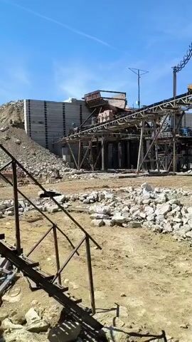 The video shows a view of a quarry or construction site with heavy machinery and piles of rocks. In the background, a large structure with conveyor belts is visible, suggesting a processing plant. A person can be seen near the machinery. The ground is covered with dirt and scattered rocks. The sky is clear and blue. The camera is positioned low, looking up towards the machinery.