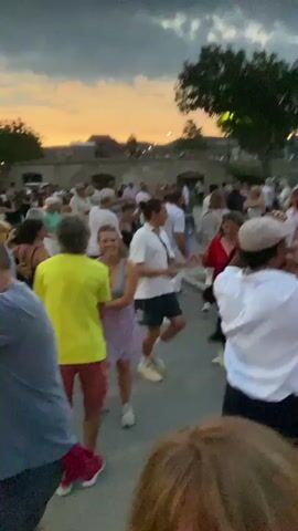 A group of people are dancing outdoors in a public space. The sky is dusky with hints of orange and yellow from a sunset. Trees and some buildings are visible in the background. Most of the people appear to be adults, dressed in casual clothing. Some are dancing in pairs, while others are dancing individually or in small groups. The overall atmosphere seems festive and relaxed.