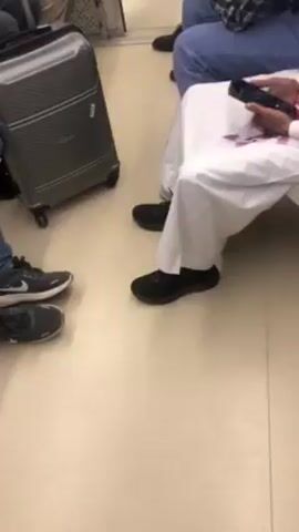 The video shows the lower portion of several people sitting in what appears to be a train car. A gray suitcase is on the floor next to a pair of sneakers. A person is wearing a traditional white thobe and black athletic shoes. They are holding a phone. Another person is wearing jeans and a plaid shirt. The floor is a light tan color.