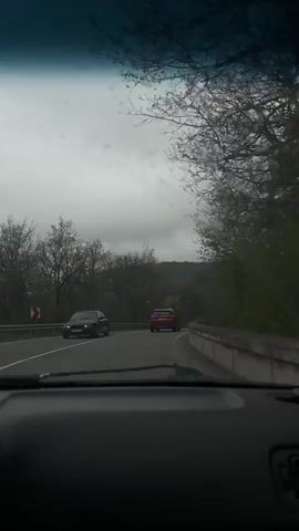 The video captures a first-person perspective from inside a moving vehicle, looking out through a rain-splattered windshield. Raindrops are clearly visible across the glass, indicating wet or recently wet weather.

At the bottom of the frame, a dark-colored dashboard is partially visible. The vehicle is traveling on an asphalt road with clear white lane markings, including a solid center line and a white shoulder line on the right. The road appears to curve gently to the right.

Ahead, a red SUV is visible, followed by another dark-colored car, both moving in the same direction. On the left side of the road, another dark-colored car can be seen, either parked or traveling in the opposite direction.

The surrounding landscape features rolling hills and mountains covered in a mix of trees. Many deciduous trees are bare or just beginning to show signs of budding, suggesting early spring. A concrete barrier or guardrail runs along the right side of the road, indicating a raised section or a bridge. The sky is overcast and gray, creating a diffused, natural light.

The primary activity is driving through a rural, mountainous environment. Dramatic, orchestral music is audible throughout the clip. There is no visible text in the video. The scene is consistent with a drive through a mountainous region, as suggested by the location context of Kostandovo, Bulgaria.