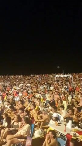 This is a vertical, shaky, low-light video of a large crowd of people, mostly young adults, seated on a beach at night. They appear to be watching something projected onto a screen, as suggested by their forward-facing gaze and the presence of what appears to be a tent or large structure in the distance, possibly where a screen might be located. The people are casually dressed, many in shorts and swimwear, and some are holding drinks or snacks. The atmosphere seems festive, with some individuals raising their arms and appearing excited. The background is dark, indicating it's nighttime, with a few faint lights visible in the distance. The camera is held at a low angle, capturing the faces and upper bodies of the people in the foreground and a densely packed crowd stretching back into the darkness.