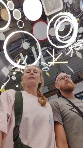 Two individuals, a woman and a man, are standing in a store aisle filled with lighting fixtures. The woman, positioned slightly in front, has blonde hair partially tied back. She is wearing a light pink t-shirt and a green shoulder bag. The man is wearing glasses, a beard, and a gray t-shirt with a black shoulder bag. The camera, held by the woman, is pointed upward at the ceiling, which is densely populated with various types of lights, including round, square, and ring-shaped fixtures. The aisle extends into the background, showing shelves with more items and other shoppers. The woman appears to be speaking, while the man stands beside her, also looking up at the lights.