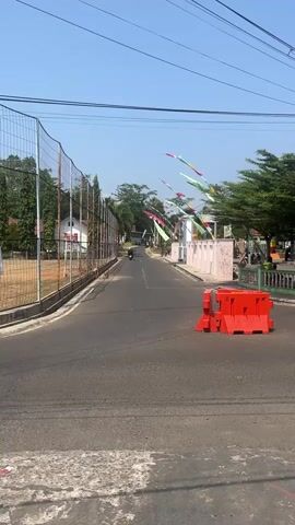 A motorcycle carrying two people rides down a paved road. To the left of the road is a chain-link fence surrounding a field. To the right of the road, there is a barricade made of orange plastic blocks. Further down the road, there are several flags or banners hanging. The sky is clear and blue, and trees line the sides of the road.