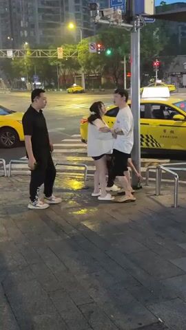 Four people are standing on a sidewalk near a street with yellow taxis and traffic lights. Two men, one in a black shirt and pants, and another in a white t-shirt and shorts, are facing a woman in a light-colored outfit. The man in the black shirt is holding a green bottle. The man in the white t-shirt appears to be talking to the woman. A fourth person, obscured by the other three, is also present. The scene appears to be at dusk or in the evening.