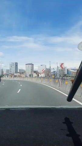 The video shows a view from inside a vehicle traveling on a highway. The highway has multiple lanes and is divided by a concrete barrier with yellow markers. Buildings of various heights are visible in the background, along with some construction cranes. Cars are also present on the highway, some in front of the vehicle and some in the opposite lanes. The sky is blue with some white clouds. A part of the vehicle's dashboard and windshield wiper are visible in the foreground. The video is approximately 5 seconds long and appears to be recorded during the daytime.
