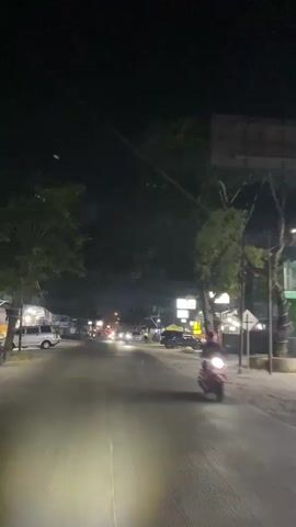 The video captures a nighttime scene on a paved road, likely in a town or city. A vehicle is moving forward, as indicated by the headlights illuminating the road ahead and the trees flanking the sides of the street. On the right side of the road, there are buildings with illuminated signs, one of which appears to be a business. Several other vehicles, including motorcycles and cars, are visible on the road, some of which are also illuminated by their headlights. The overall atmosphere is that of a suburban or urban street at night, with ongoing traffic and illuminated businesses.