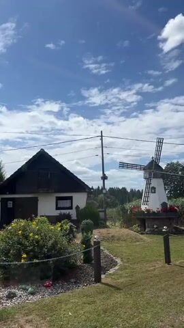 The video begins with a view of a white windmill with black blades against a blue sky with scattered clouds. A white building with a dark roof is visible to the left of the windmill. A metal pole with wires runs across the frame in front of the windmill. In front of the windmill, there is a stone base adorned with red flowers. A grassy area with two white-capped posts and a chain fence separates the viewer from the windmill. The video then pans upward to focus on the sky and clouds.