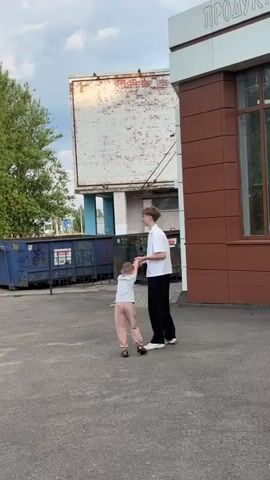 A young male wearing a white shirt and black pants stands on asphalt, holding the hands of a younger child who is wearing a light-colored top and pink pants. The male spins the child around. In the background, there are dumpsters and a building with a brown facade. The sky is overcast.