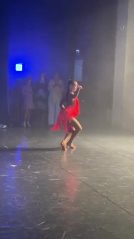 A girl in a red fringed dress dances on a stage in front of an audience. A boy in a white shirt and black pants joins her. They perform a synchronized dance. The stage is dimly lit with blue spotlights.
