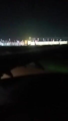 The video shows a view from an airplane window at night. The wing of the plane is visible in the foreground, obscuring most of the view below. In the distance, there are lights that appear to be from buildings, vehicles, or streetlights on the ground. The lights are blurry, suggesting motion or distance. The sky is dark. The scene is dim overall, making it difficult to discern details.