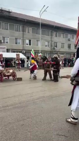 The video shows a group of people dressed in traditional Bulgarian costumes performing a ritual in a town square. The costumes are elaborate, with large masks and colorful fabrics. Some of the performers are carrying wooden sticks, while others have bells attached to their belts. There is also a small wooden cart on display. The scene is festive and lively, with music playing and people moving around. In the background, there is a building with several windows and a few trees. The overall atmosphere is one of cultural celebration.