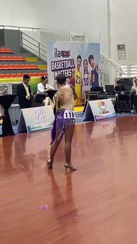A young woman in a sparkly purple and gold dance costume with a number 110 on her back performs a dance routine on a polished wooden floor indoors. She has her arms raised and is looking upwards. Behind her, a large banner displays "ILA Basketball Masters" with cartoon characters and text in English. Several people are seated or standing behind the banner, some of whom appear to be judges or officials. In the background, orange bleachers are visible. The lighting suggests an indoor sports arena or gymnasium.