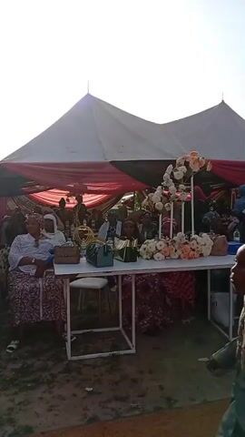 The video shows an outdoor event, possibly a wedding or celebration. A white tent with red and black lining covers tables decorated with flowers. People are seated at the tables, and some are standing and dancing. Many women are wearing colorful, traditional African clothing and headwraps. One woman is carrying a baby. The scene is lively, with people interacting and enjoying themselves. The weather appears sunny.