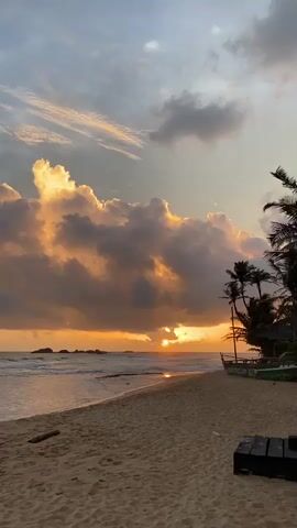 This is a landscape video of a beach scene at sunset in Hikkaduwa, Sri Lanka. The main subjects are the natural elements of the beach, ocean, and sky. The scene depicts a sandy beach with a gentle shoreline where waves are rolling in. In the distance, there are several rock formations jutting out of the water.

The sky is filled with dramatic clouds, illuminated by the setting sun which is partially obscured by them, casting a warm, golden glow. The clouds themselves range in color from deep grey to bright orange and yellow where the sunlight hits them. Palm trees line the edge of the beach on the right side, with a small, traditional-looking boat resting on the sand. There is also a dark, rectangular object that appears to be a piece of furniture or equipment on the sand in the foreground.

The time of day is clearly sunset, evidenced by the low sun and the warm, diffused light. The weather appears to be partly cloudy, creating a visually striking sunset. The location is identifiable as a tropical beach with palm trees and ocean views. The overall emotion evoked by the scene is one of tranquility and natural beauty. There is no visible text in the media file.
