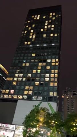 The video pans across a cityscape at night, featuring tall buildings with illuminated windows. One building has a unique design with angled sections. Trees in the foreground are adorned with decorative lights, some featuring purple flowers. A woman is seen standing near the trees, looking at her phone. The sky is dark, suggesting a late evening or nighttime setting.