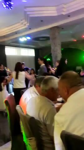 The video shows a lively indoor gathering, likely a celebration or party, with several people seated at tables and others dancing. The environment appears to be a restaurant or banquet hall, featuring a decorative ceiling with colored lights, a large stone-like pillar, and chandeliers. In the foreground, several individuals are seated at a table, facing away from the camera, engrossed in conversation or the event. Some are wearing casual attire, while others are in dressier clothes. The middle ground and background reveal more activity, with a group of people, including women and children, dancing and socializing. The lighting is dim and colorful, with flashing disco lights adding to the festive atmosphere. There are bottles and food on the tables, suggesting a meal is being served. The overall impression is one of a joyful and energetic social occasion.