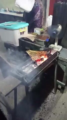 The video shows a person grilling satay over charcoal. The grill is a metal rectangular box with a grate. Satay skewers are arranged on the grate, cooking over glowing coals. Smoke rises from the grill. To the left of the grill, there is a metal table with a black container. Behind the grill, there is a white and teal plastic container. A white fan is visible to the right of the grill. In the background, there are tables and chairs, suggesting a food stall or small restaurant setting. A person in a patterned shirt is partially visible standing behind the grill. The floor appears to be concrete, and the walls are a neutral color. The lighting is dim, indicating it might be evening or indoors.
