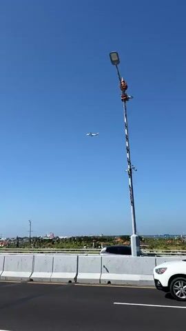 A white car drives on a road. A plane flies in the sky above. The sky is clear and blue. There are some buildings in the distance. There is also a lamp post on the side of the road.