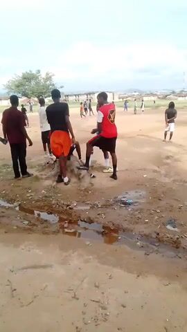 The video shows a group of young men standing on a dirt field with puddles of water and debris. Some are wearing athletic clothing, including shorts and jerseys. One person in a red and white jersey appears to be kicking or nudging something on the ground, while others are observing or interacting with each other. In the background, there are more people and structures that suggest an outdoor gathering or informal sporting activity. The sky is cloudy.