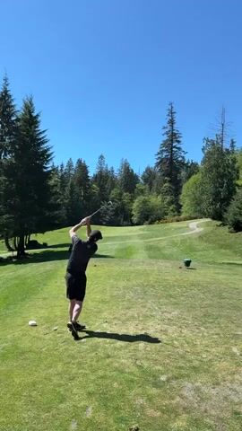 A man dressed in a black t-shirt and black shorts is playing golf on a bright, clear day. He has just swung his golf club, hitting the ball toward the fairway. He is standing on the tee box of a green golf course, with tall trees visible in the background. The sky is clear and blue.