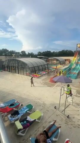 The video shows a water park on a cloudy day. In the foreground, several lounge chairs are set up on a concrete patio. One chair in the lower right corner has red and white striped items on it. In the mid-ground, a lifeguard sits in a raised chair wearing a yellow vest and a whistle around their neck. Behind the lifeguard are two large, brightly colored water slides, one green and blue, the other orange and yellow. People are visible on the slides and in the pool at the bottom of the slides. To the left of the slides is a covered pool area with more lounge chairs. In the background, there are trees and several small cabins. The sky is a mix of blue and white clouds. The overall scene depicts a busy water park with various attractions and people enjoying their time.