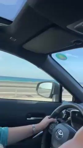 A person is driving a Toyota on a highway with the ocean visible in the distance. The driver has a watch on their left wrist. The car's dashboard displays a navigation system. There are other vehicles on the road. The sky is blue and sunny. The interior of the car is black. There is music playing. The car has a sunroof.
