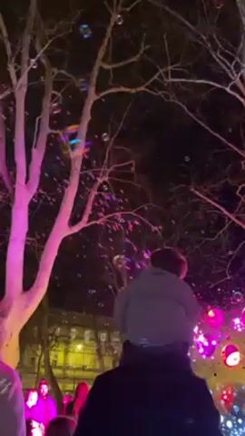 At night, an outdoor scene features a crowd of people enjoying a bubble show. The bubble show is characterized by a multitude of bubbles floating in the air, illuminated by bright, colorful lights ranging from purple to yellow. Bare tree branches are visible against the dark sky, with some of the branches also illuminated by the lighting. The individuals in the crowd appear to be looking up at the bubbles, and their faces are partially visible in the light. The overall atmosphere appears festive and celebratory.