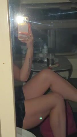 The video shows a woman sitting in what appears to be a train compartment. She is wearing a dark dress and taking a selfie with her phone in front of a mirror. The train compartment has a small table with a water bottle and other items on it, and a bed. The woman has a tattoo on her arm. The lighting is dim inside the train compartment.