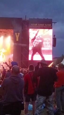 The video shows a concert with a stage and a large screen displaying performers. Many people are gathered in front of the stage, some of whom are filming the event. The screen shows a person playing a guitar. The sky is dark, suggesting it is evening or night. The concert appears to be a rock or metal concert, judging by the stage lighting and the attire of some of the concertgoers.