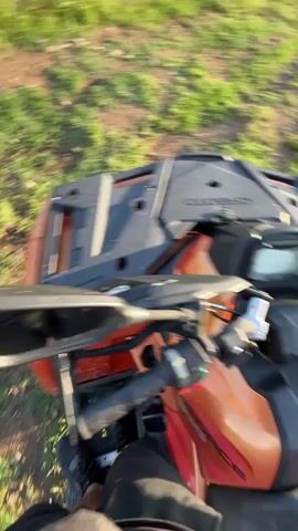 The video captures scenes from an outdoor, rural setting, likely in Anevo, Bulgaria, during the late afternoon or early evening under a bright, sunny sky with scattered clouds.

The initial shots provide a first-person perspective from an All-Terrain Vehicle (ATV), showing its dashboard with "CFMOTO" partially visible, handlebars, and a blurry green foreground as if in motion. Subsequent POV shots from the ATV reveal a vast landscape. A dirt road winds through green fields, leading towards distant hills and mountains under a bright sun.

The video then shifts to a view of a dirt track, possibly an off-road or rally course, bordered by a fence made of concrete posts and stones. This track is situated amidst green fields, with the mountains forming a scenic backdrop.

Finally, the scene shows two adult men and a small child standing near the fence, observing the dirt track and the expansive landscape. One man wears a blue hoodie and shorts, while the other wears a black t-shirt, shorts or pants, and carries a shoulder bag. The child is dressed in light-colored clothing. They appear to be enjoying the view and the outdoor environment.
