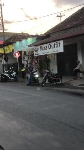 The video shows a street scene at dusk. Several motorcycles are parked along the side of the road in front of shops. One motorcycle in the foreground has a rider on it, appearing to be in motion. A person is sitting on a bench outside one of the shops. The sky is overcast with some light visible. The shops have signage, one of which reads "Mira Outfit".