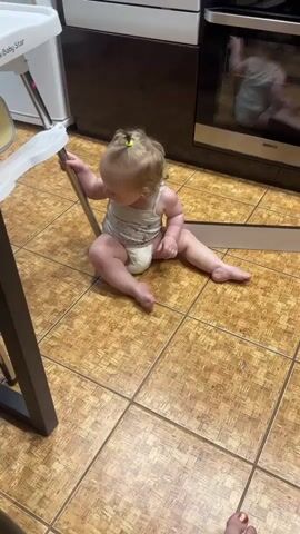 A baby is sitting on the floor in a kitchen. The baby is wearing a diaper and a tank top. The baby is leaning on a high chair and a stove. The baby is looking at the stove. The floor is tiled. There is a refrigerator in the background.
