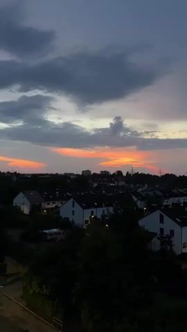 The video shows a sunset over a residential area. The sky is filled with clouds in shades of gray, blue, and pink, with the sun creating a vibrant orange glow on the horizon. Below the sky, a row of houses with white walls and dark roofs is visible, nestled among dark green trees. The houses are illuminated by the fading light, and some lights are on inside. The overall atmosphere is calm and serene.