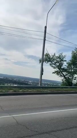 This video captures a view from a moving vehicle on a road. The road surface is asphalt with white lane markings. To the left, there is a metal guardrail bordering a slope leading down to a valley. In the valley, a town or village with scattered houses and trees is visible. A tall utility pole with wires stretches across the frame in the foreground. A single tree with green leaves is positioned to the right of the utility pole. The sky above is partly cloudy with a mix of blue sky and white clouds. At one point, a light-colored car is seen driving on the road ahead. The overall environment appears to be rural or suburban, with a landscape of rolling hills and vegetation under a daytime sky.