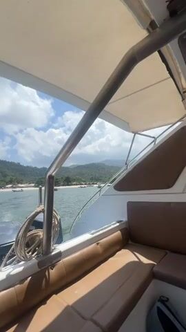The video captures a view from the back of a boat, looking out towards a tropical coastline. The boat is equipped with two Yamaha 200 horsepower outboard motors, which are actively propelling the vessel, creating a wake behind it. Brown cushioned seating is visible in the foreground, along with metal railings and a rope coiled on the deck.

In the background, a sandy beach is visible, lined with dense green trees and lush mountains or hills under a bright blue sky dotted with fluffy white clouds. The water is calm and clear, with a few other smaller boats anchored or moving near the shore. The scene suggests a sunny daytime setting, likely a leisurely boat trip around the coast of Koh Phangan, Thailand, as provided by the context.