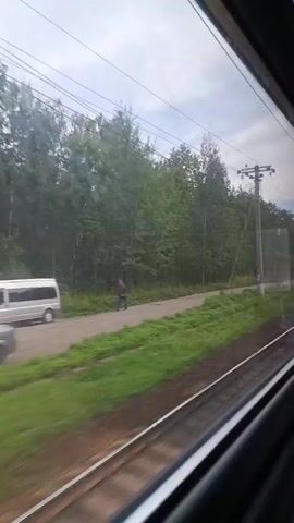 The video shows a view from a moving train, looking out the window at a rural or semi-rural landscape. The train tracks are visible in the foreground, with green vegetation bordering them. Further out, there are trees and power lines, typical of a train route. In the distance, a road runs parallel to the tracks, and a few vehicles, including a silver car and some construction equipment, are visible. The sky is overcast. The camera is panning slightly, suggesting the train is in motion. The scene appears to be an ordinary day in a temperate climate. There are no people clearly visible or interacting with each other.