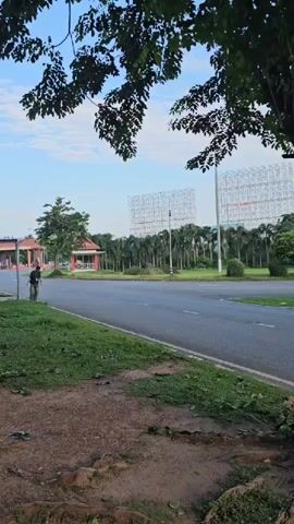 The video begins with a wide shot of a street with a person on a bicycle in the distance. The camera then pans to a group of people sitting on the side of the road, some eating. It then shifts to a person standing under a shelter, followed by a shot of a person sitting on a bench. The video concludes with a shot of a building with people sitting on benches outside. The environment appears to be a park or a public area with trees and buildings visible.