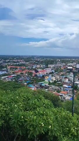 The video captures an aerial view of a sprawling city, seemingly taken from a hilltop. The foreground is dominated by lush green foliage, suggesting a natural vantage point overlooking the urban landscape. In the mid-ground, a light pole with a rectangular light fixture stands prominently, a man-made element amidst the greenery. Beyond this, the city unfolds with a dense collection of buildings of varying colors and sizes, exhibiting a mix of residential and commercial structures. The architecture appears to be predominantly of Asian design, with tiled roofs in shades of orange and red. The urban sprawl extends to the horizon, where it meets a hazy sky filled with dramatic, gray clouds. The overall impression is one of a vibrant, densely populated city viewed from a distance, with a blend of natural and man-made elements creating a picturesque panorama. The light and cloud formations suggest a potentially overcast or stormy weather condition.
