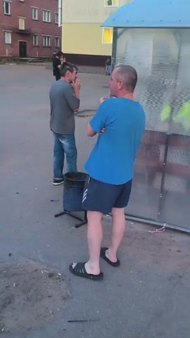 A man in a blue t-shirt and dark shorts stands facing away from the camera, looking towards a transparent shelter with a blue roof. He is wearing black flip-flops. To his left, another man in a grey t-shirt and blue jeans stands facing him and also looking towards the shelter. He is holding something to his face, possibly a cigarette. A black trash can with a metal stand is situated between the two men. In the background, a brick apartment building and a yellow building are visible. Other figures are present in the distance. The scene appears to be outdoors in what looks like a courtyard or residential area.