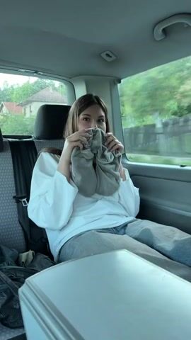 Here is a detailed description of the video:

The video shows a young woman in the back seat of a moving vehicle. She is wearing a white long-sleeved shirt and gray pants, and is buckled in with a seatbelt. She is holding a gray travel pillow up to her face, covering her nose and mouth. She looks at the camera and smiles slightly. The vehicle appears to be traveling through a rural area with trees and houses visible through the window. There is a gray table in front of her. The interior of the vehicle appears to be clean.